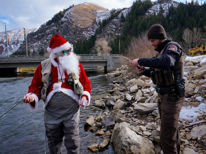 (Bethany Baker  |  The Salt Lake Tribune) Devin Shirley, right, a conservation officer with the Utah Division of Wildlife Resources, checks the fishing license for Rudy Schenk, a fly fisherman dressed as Santa Claus, in the Provo River at Vivian Park in Provo Canyon on Saturday, Dec. 23, 2023.